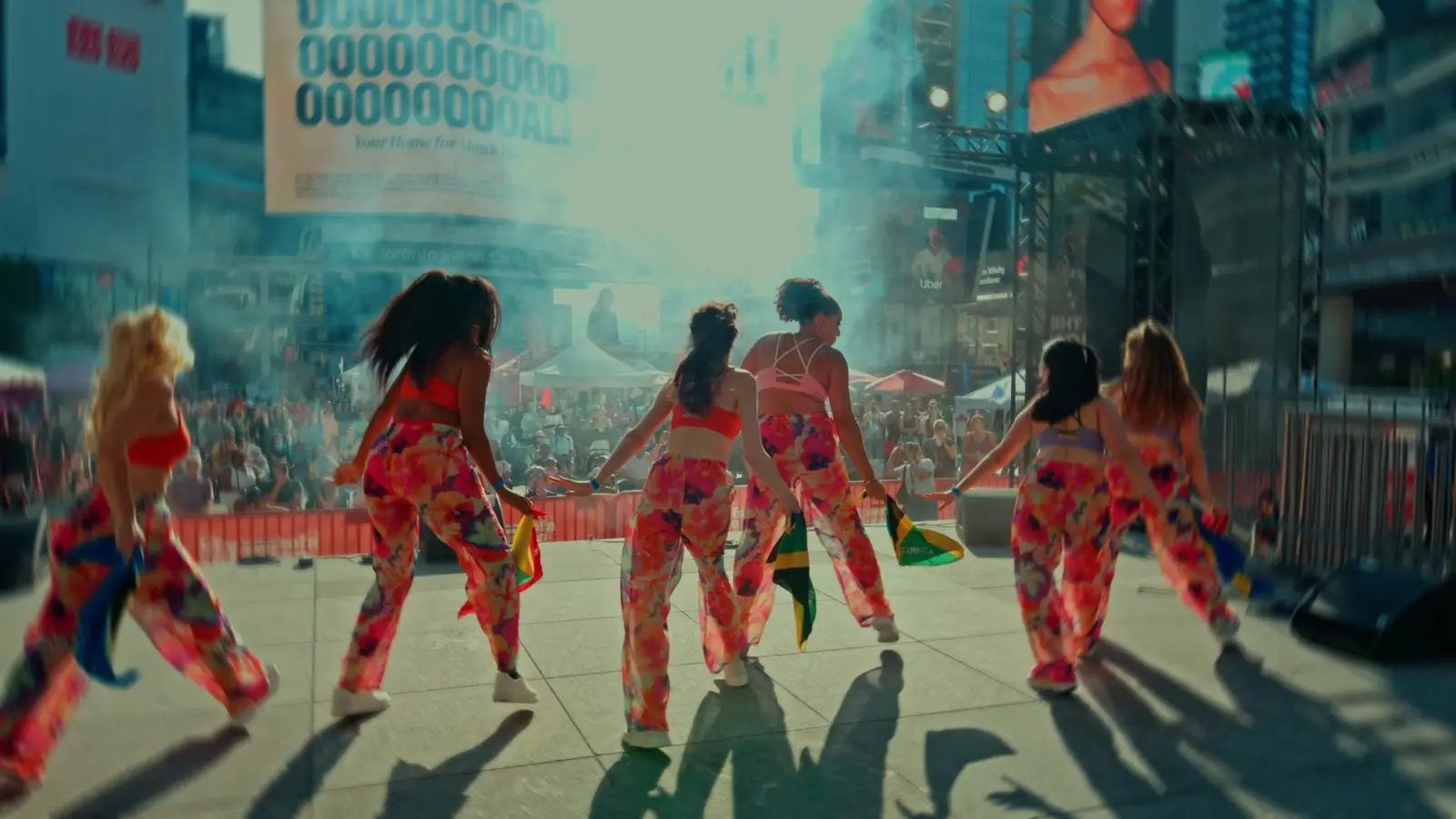 A dance troupe in bright printed outfits performs through stage smoke, facing the crowd