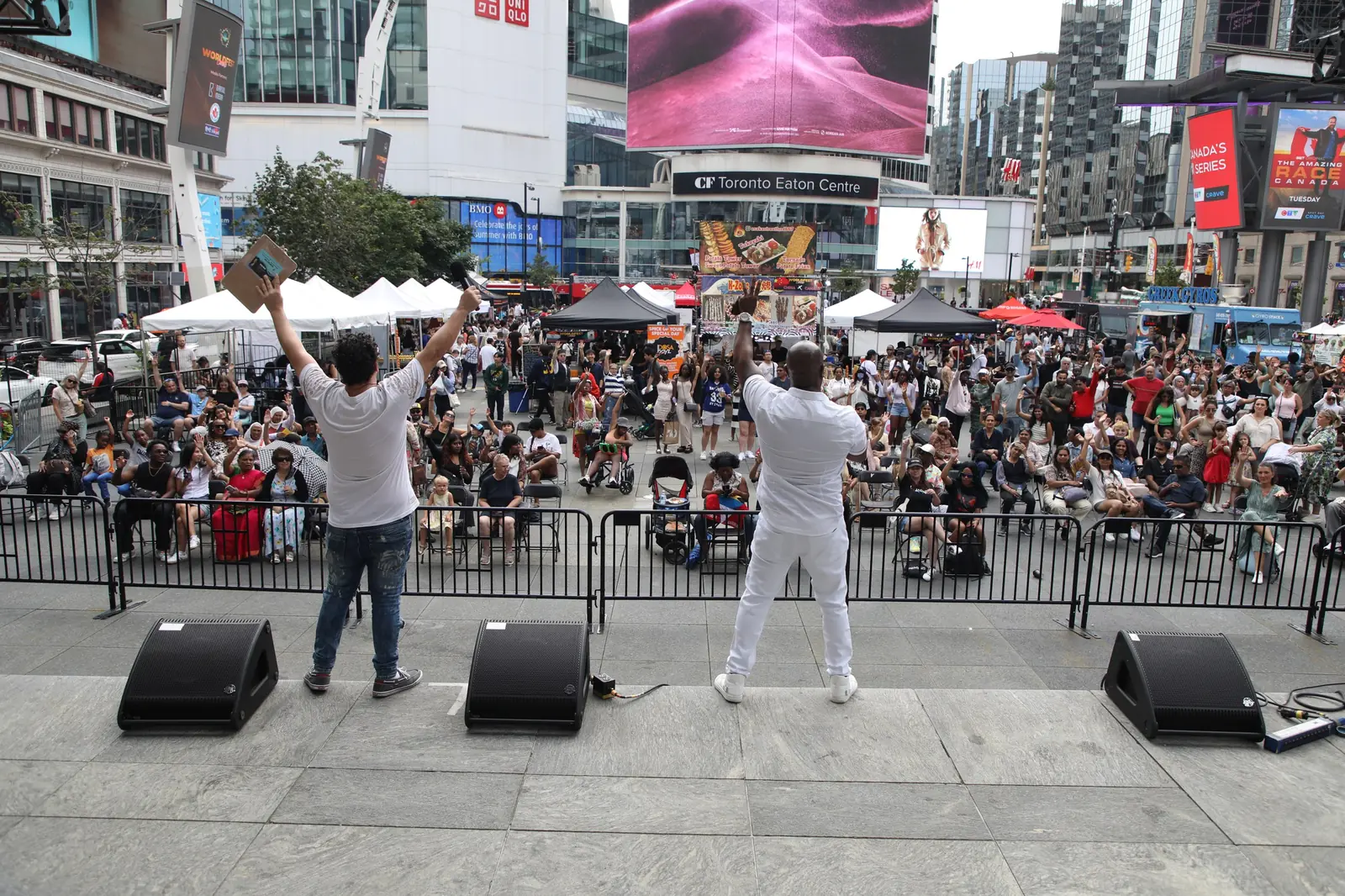 Hosts on the WorldFest main stage raise their arms to a cheering crowd at Sankofa Square, with the Toronto Eaton Centre behind